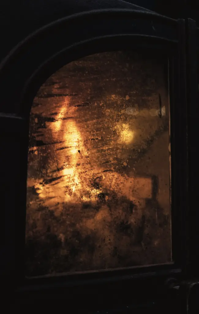 Vitre d'un poêle en fonte voilée de suie, le feu rougeoie derrière
